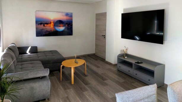 Un salon moderne avec un canapé gris, une table basse ronde en bois, un téléviseur à écran plat fixé au mur et un grand tableau abstrait.