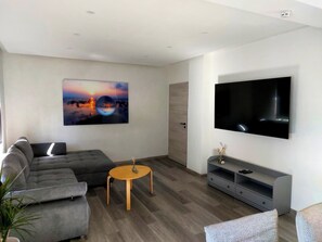 A modern living room with a grey sofa, a round wooden coffee table, a flat-screen TV mounted on the wall, and a large abstract painting.