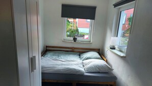 Une petite chambre avec un lit simple, une fenêtre donnant sur un immeuble rouge et une porte située à gauche.