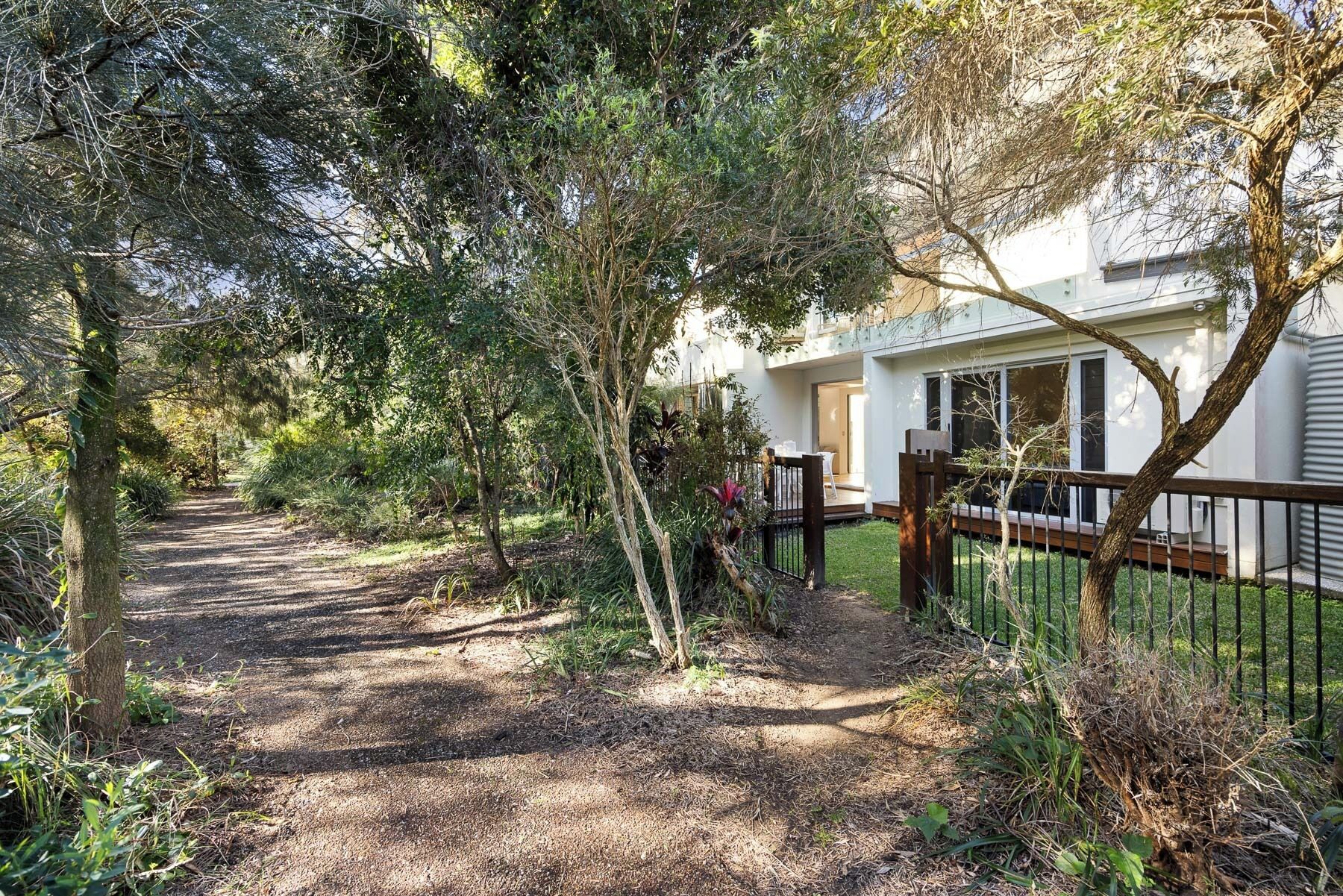Un chemin mène à un bâtiment blanc, entouré d’une clôture en bois et bordé d’une végétation luxuriante.