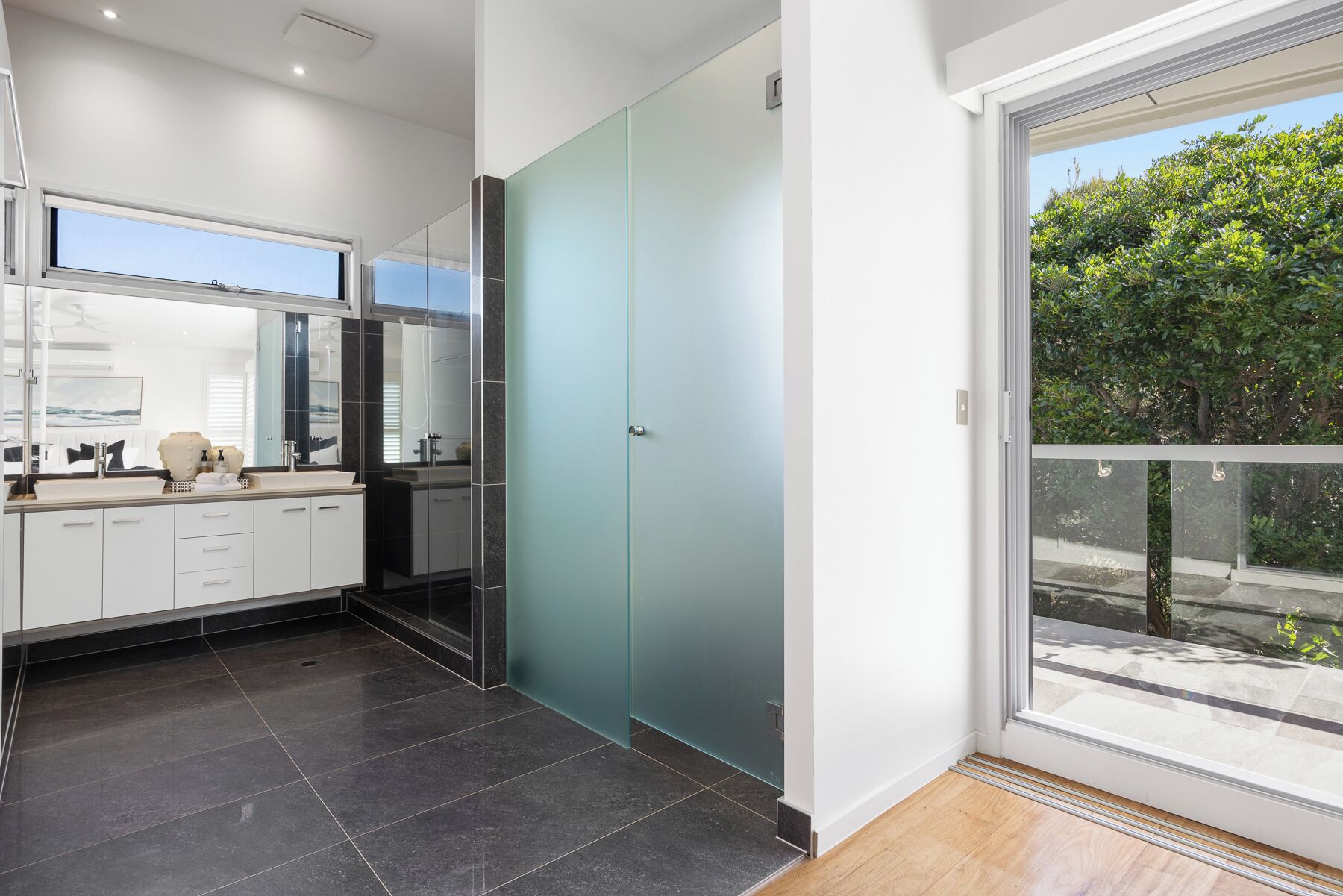 Een moderne badkamer met een douchecabine van glas, een grote spiegel en uitzicht op groen via een schuifdeur.