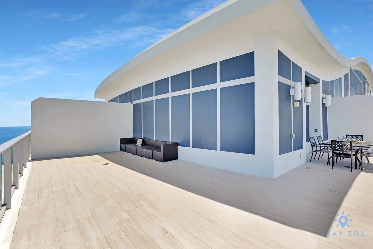 A modern outdoor deck with a seating area, large windows, and a view of the sea.