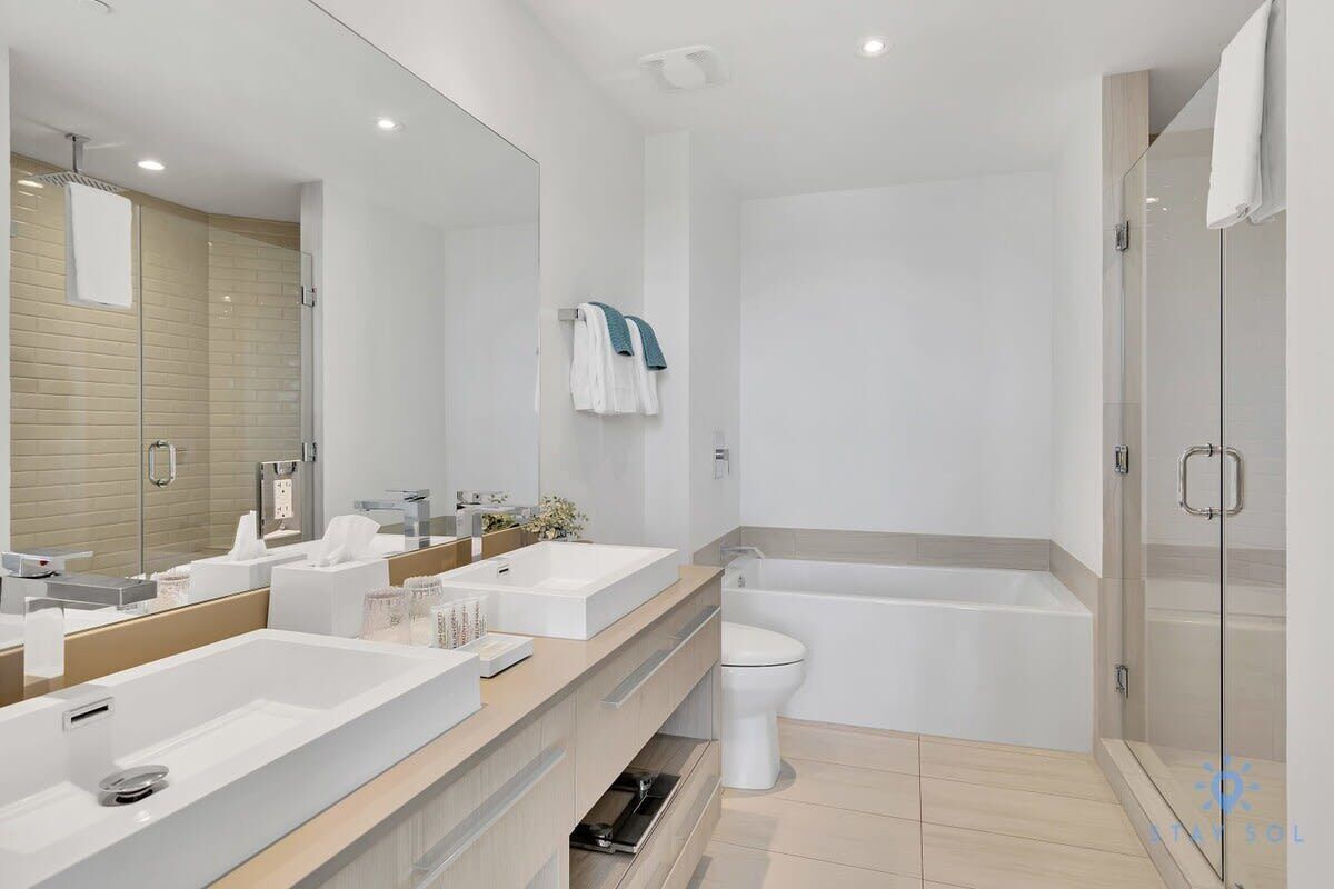 A modern bathroom with a large mirror, two sinks, a toilet, and a bathtub.
