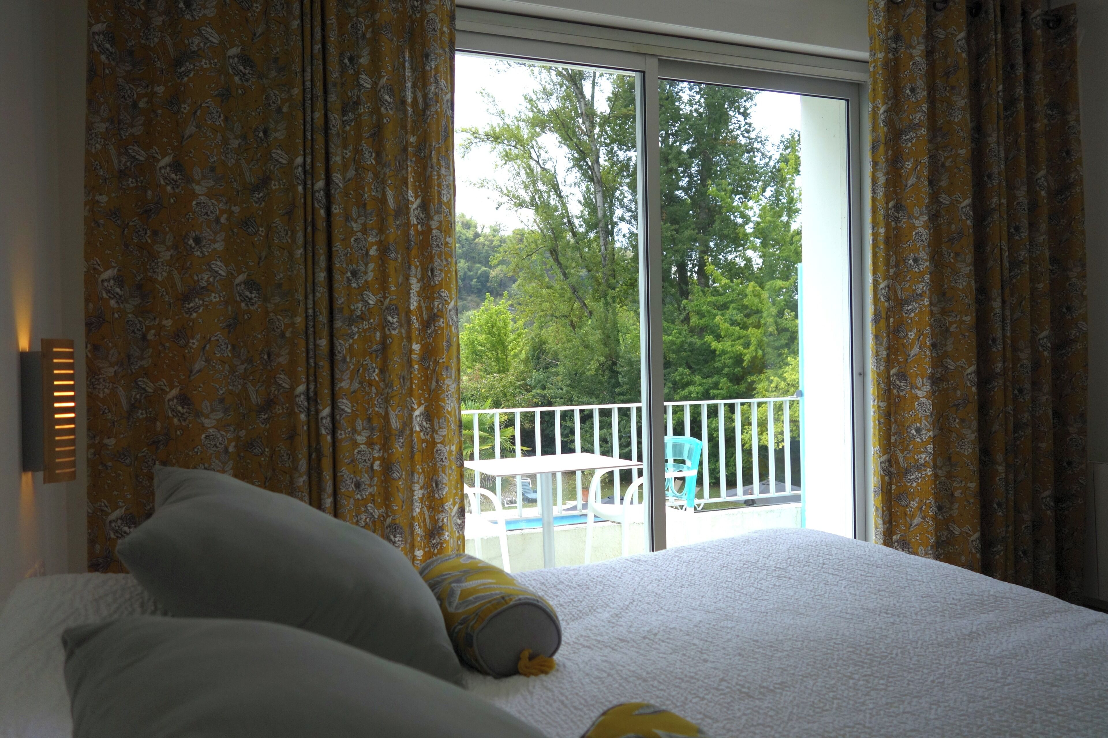 A bedroom with a large bed, floral curtains, and a view of a balcony with outdoor furniture.