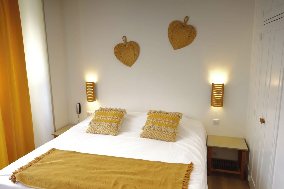 A neatly made bed with a yellow throw blanket, two patterned pillows, and two wall-mounted light fixtures.