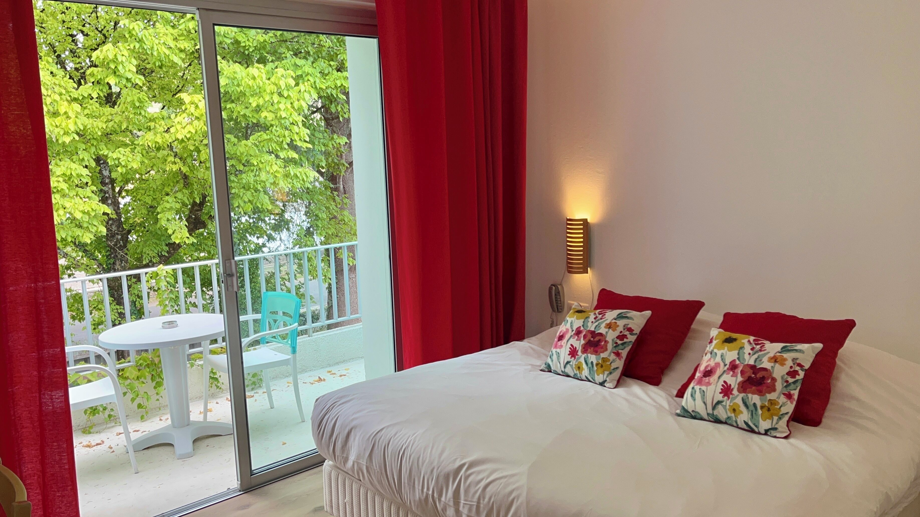 Habitación de hotel con cama, cortinas rojas, balcón con mesa y sillas, y vista a árboles.