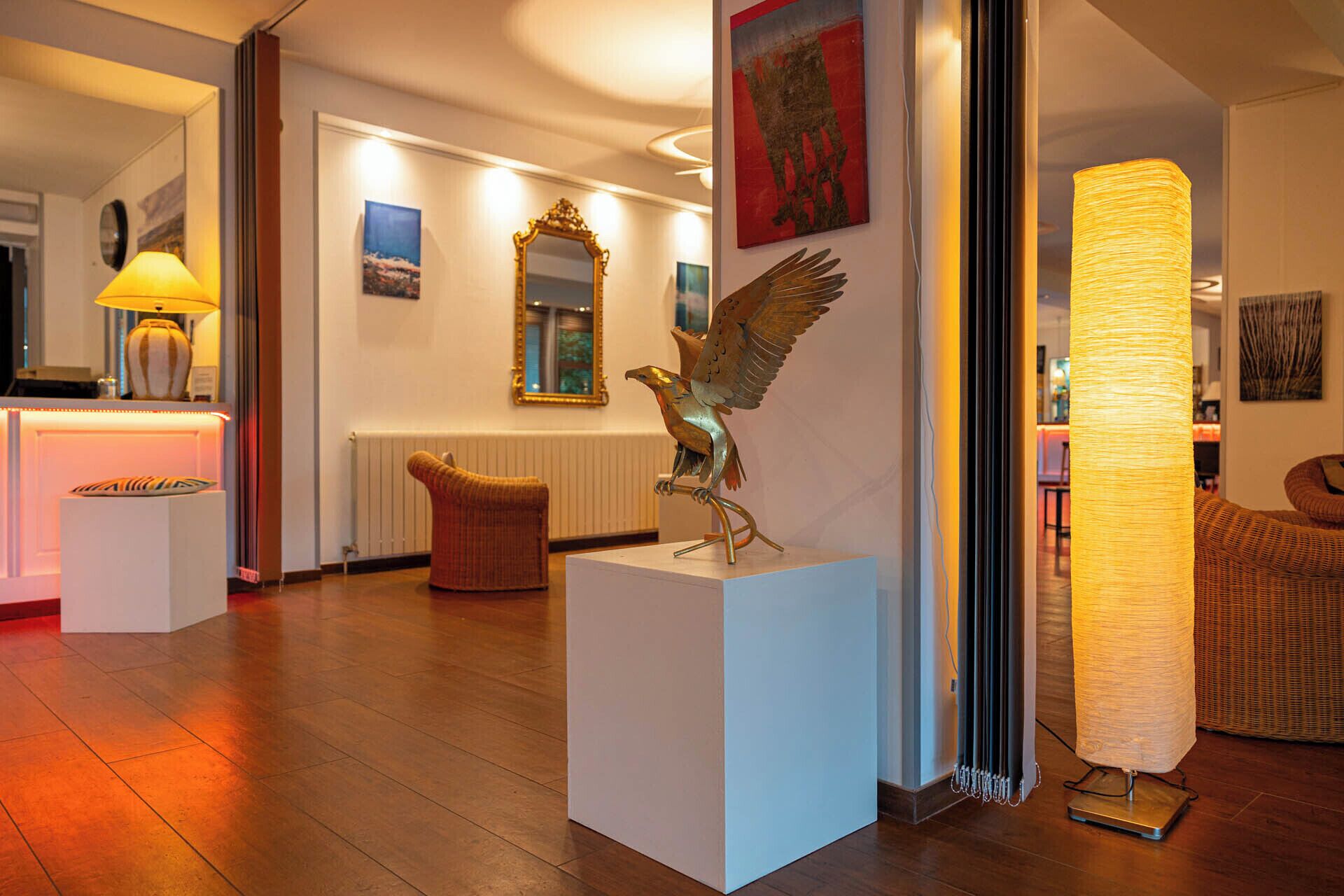 A modern interior with a wooden floor, a white reception desk, a wicker chair, a sculpture of a bird, and a tall, textured lamp.