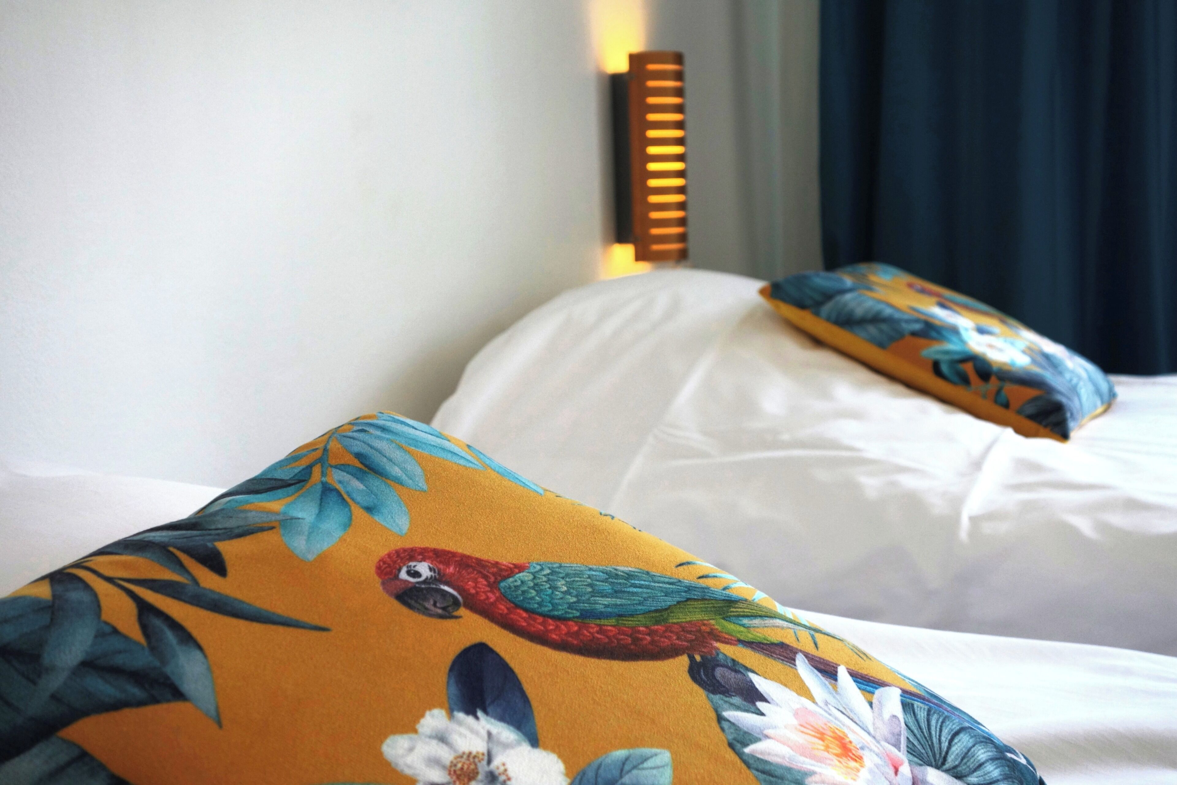 Cama con sábanas blancas y almohadas coloridas con diseño de loro y motivos florales.