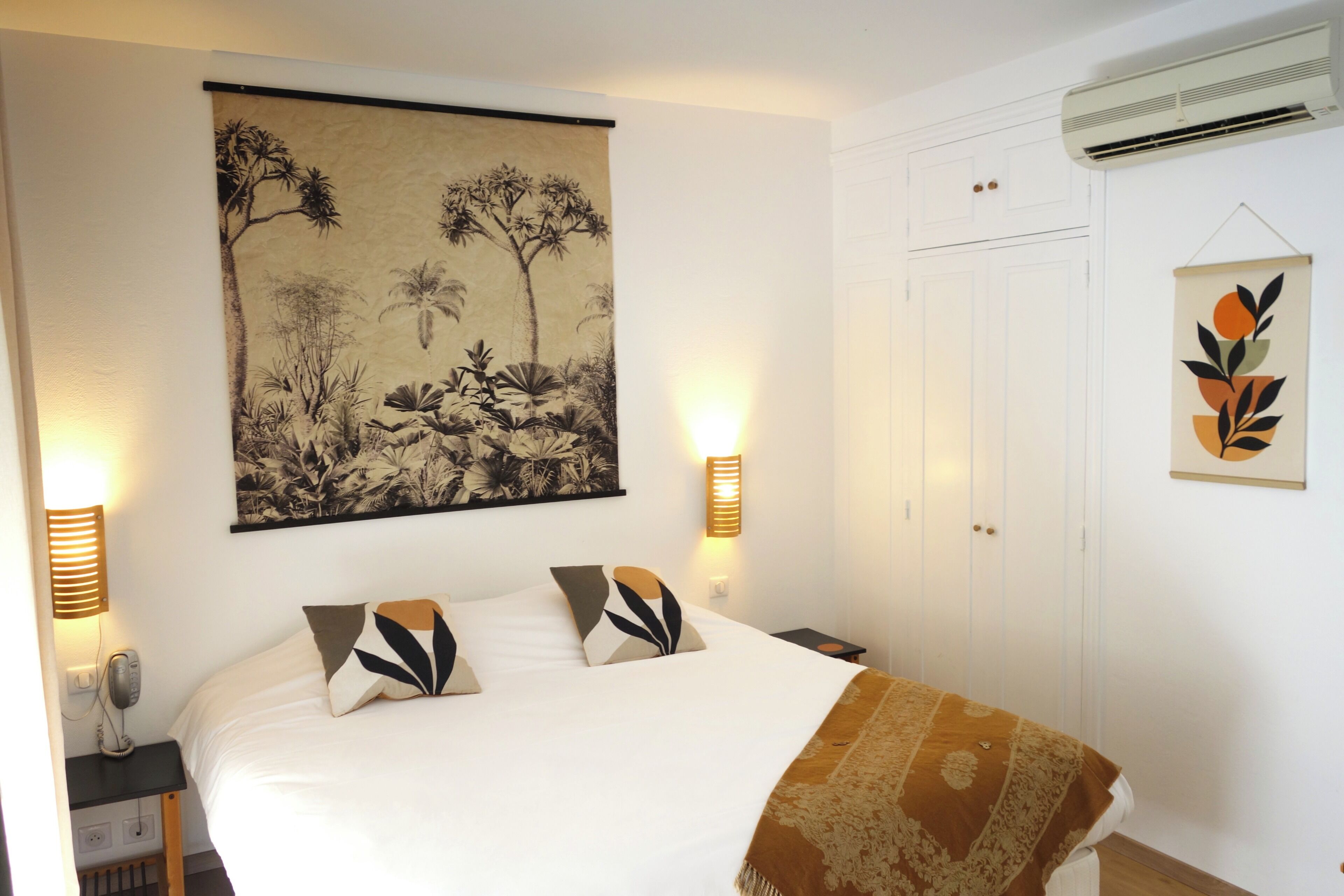 Un dormitorio con una cama grande, dos lámparas de noche, aire acondicionado de pared y una obra de arte enmarcada sobre la cama.