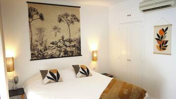 Un dormitorio con una cama grande, dos lámparas de noche, aire acondicionado de pared y una obra de arte enmarcada sobre la cama.