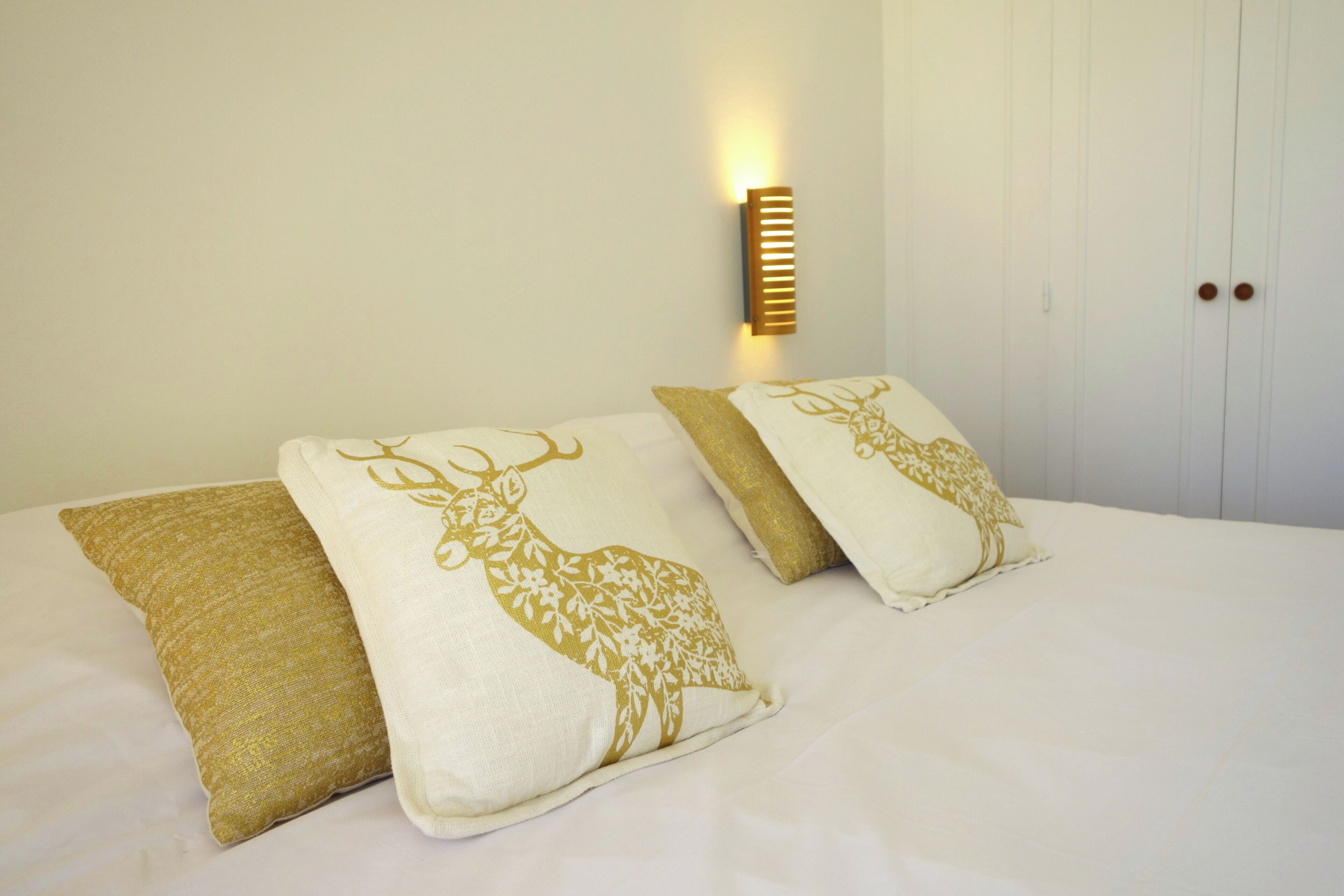 Una cama con dos almohadones decorativos con diseño de ciervo, una lámpara de pared y un armario blanco con tiradores de madera.