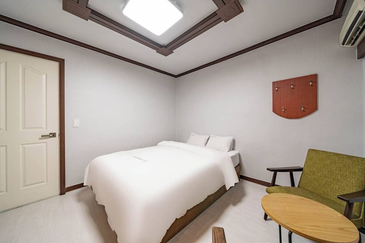 A bedroom with a bed, a green armchair, a small wooden table, and a wall-mounted decoration.