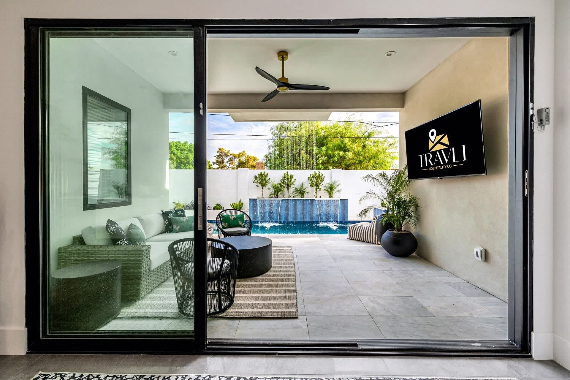 A modern hotel room with a sliding glass door leading to a pool area, a ceiling fan, and a mounted TV displaying the TRAVLI logo.