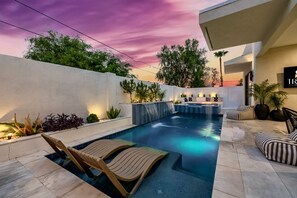 Una piscina con caída de agua, sillas de descanso y una zona de estar con vistas a la puesta de sol.