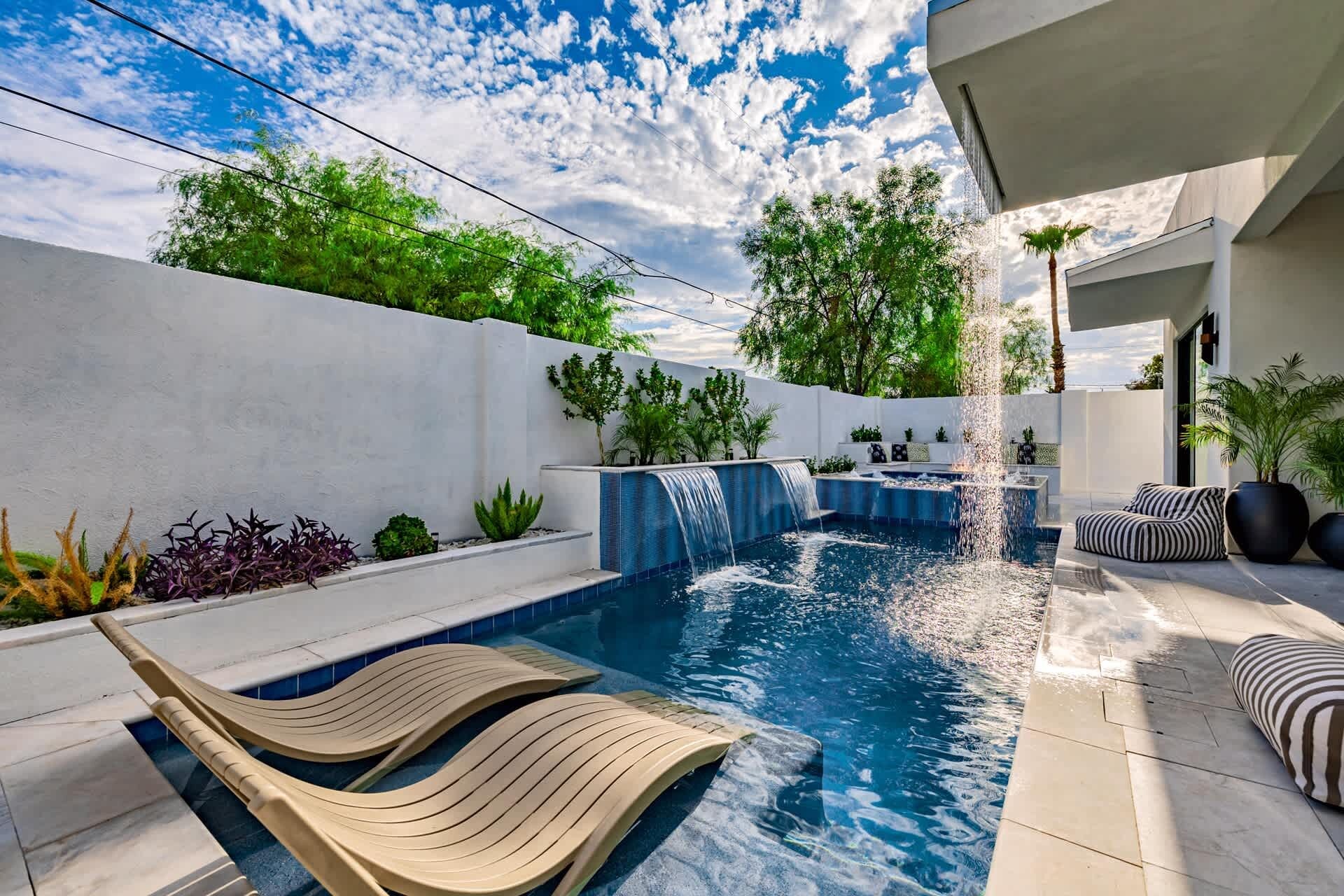 Un área de piscina moderna al aire libre con una cascada, sillas de descanso y plantas en macetas.