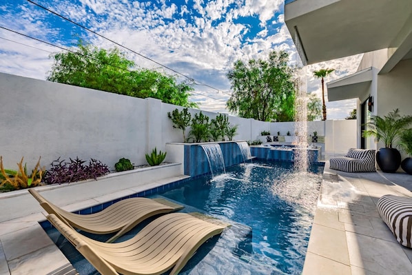 Un área de piscina moderna al aire libre con una cascada, sillas de descanso y plantas en macetas.