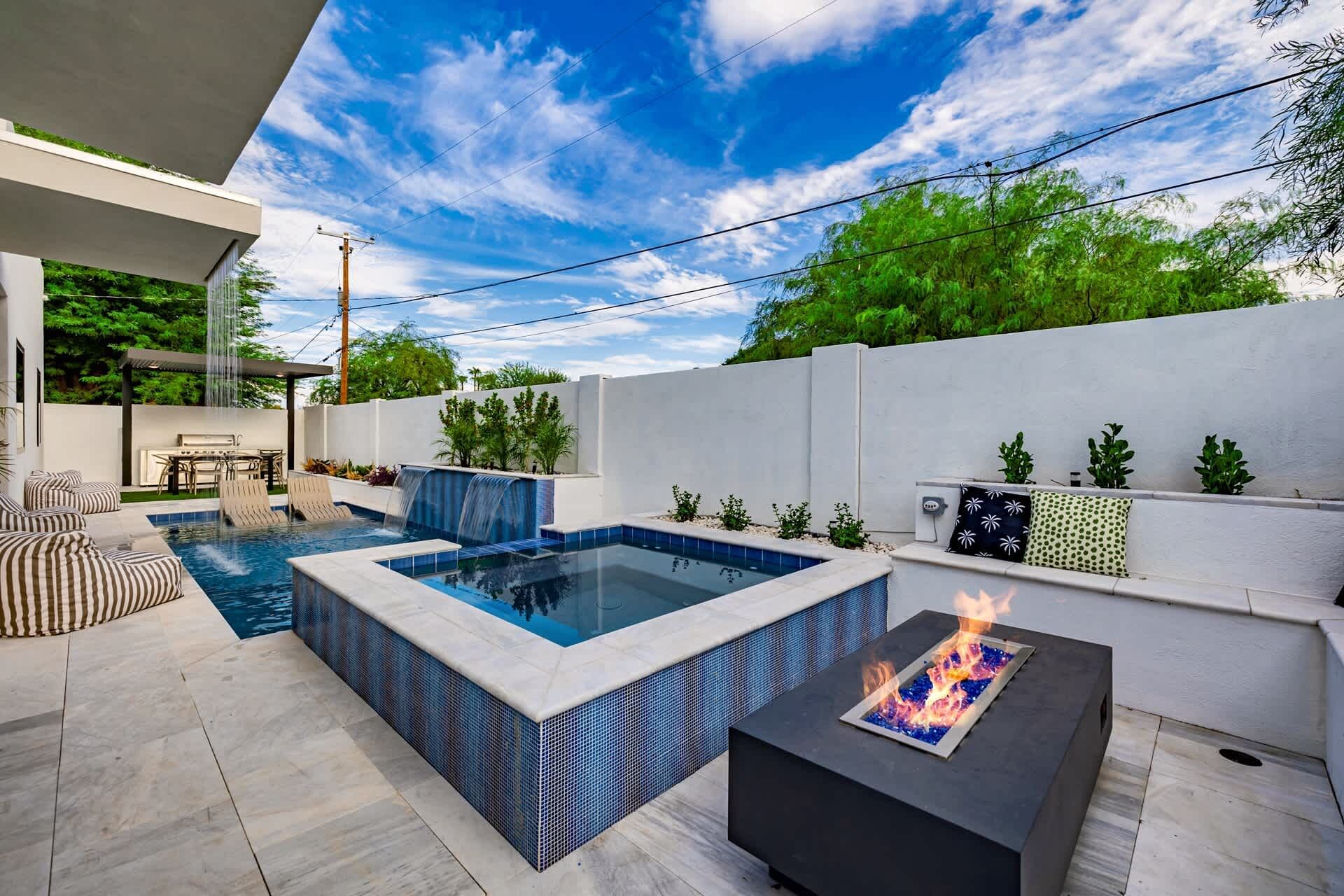 Un área de piscina exterior moderna con jacuzzi, asientos y una chimenea.