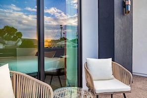 Un balcón con sillas de mimbre y una mesa de vidrio, con vistas a un paisaje urbano que incluye edificios y un cielo despejado.