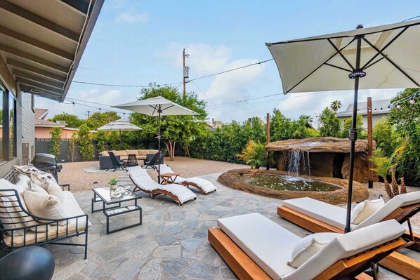 A patio with outdoor seating, a water feature, and a covered area with umbrellas. - SolaNova Retreat | Cocktail Pool & Movie Room (Scottsdale)
