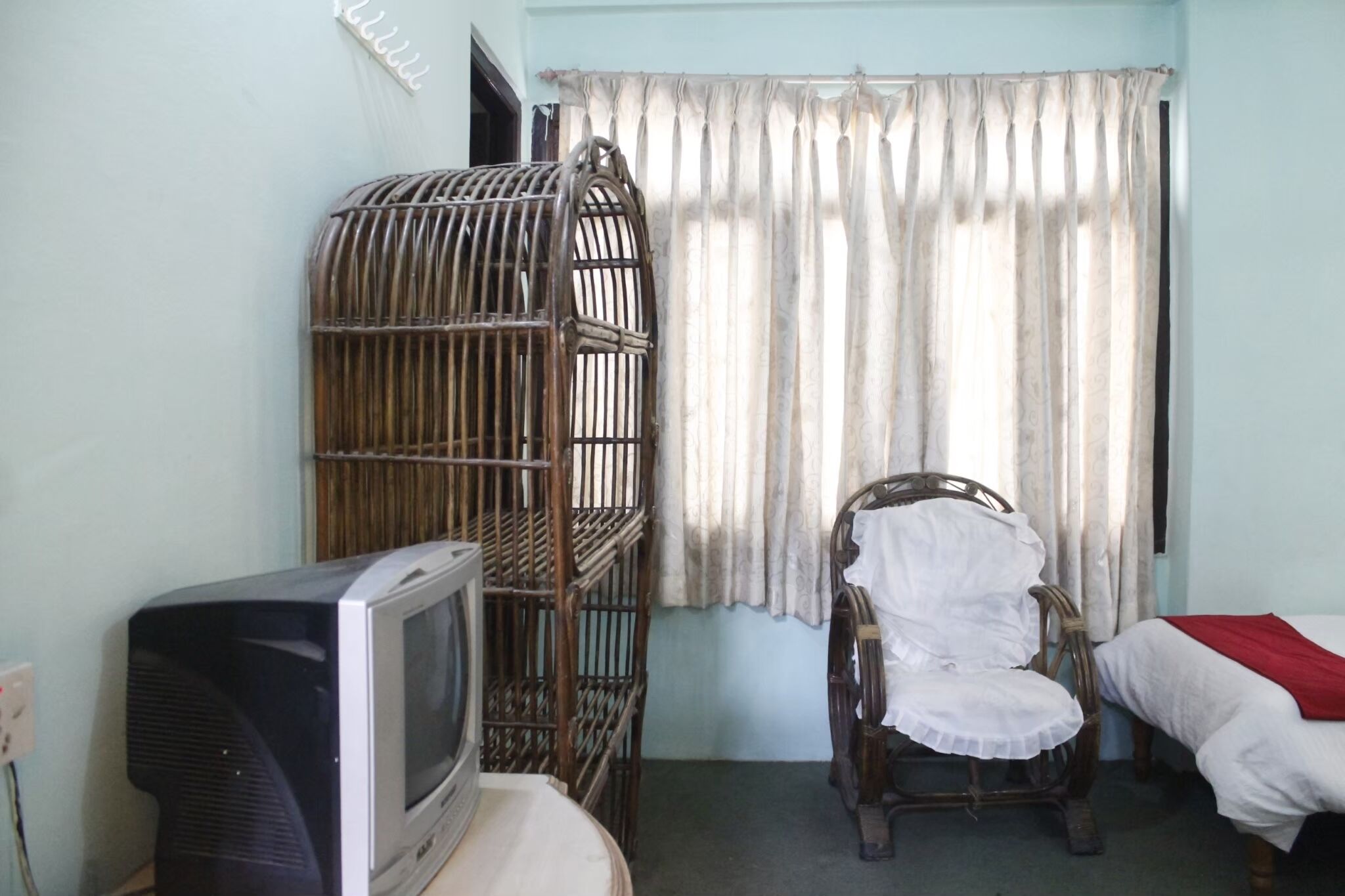 Una habitación con televisión, una silla de mimbre, una cama con una colcha roja y una ventana con cortinas.