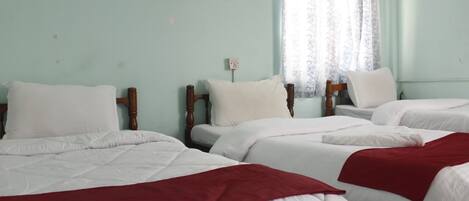 A hotel room with three beds, each with white bedding and a red blanket on the lower bed. There is a window with sheer curtains and a wall-mounted light switch.
