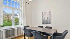 A dining area with a long table and six chairs, a radiator, a window with a view of buildings, and a framed cityscape picture on the wall. - Spacious & Tranquil Room in Limpertsberg (Luxembourg)