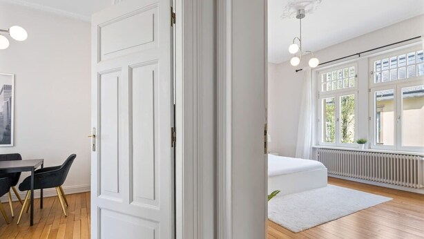 A room with a white door, a dining table with chairs, a white sofa, and a large window with a view of greenery. - Spacious & Tranquil Room in Limpertsberg (Luxembourg)