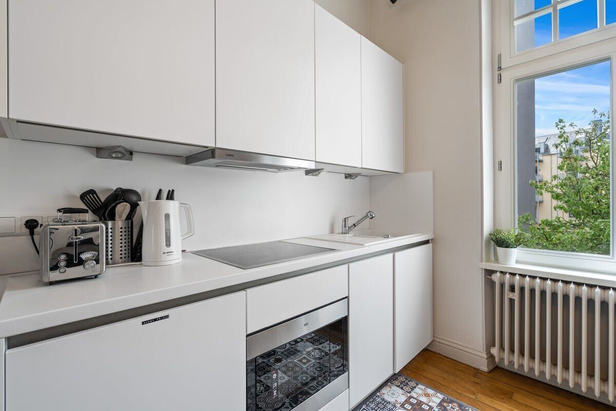 Ett modernt kök med vita skåp, en inbyggd ugn, en brödrost och ett fönster med utsikt över byggnader och grönområden.