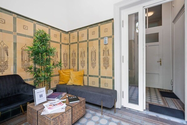 Un área de recepción con un sofá, una mesa de centro con revistas, una planta en maceta y una pared con azulejos decorativos.
