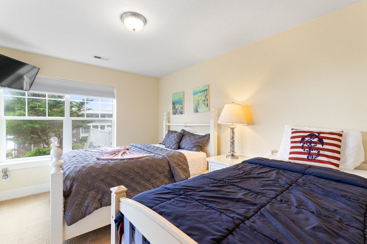 A bedroom with two beds, a window with blinds, a lamp, and artwork on the wall.