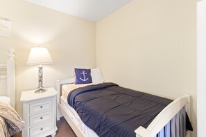Une chambre à coucher avec un lit, une table de chevet avec une lampe, un oreiller orné d’un motif d’ancre et une commode blanche.