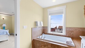 A bathroom with a large bathtub, a window with a view, and a bed in the adjacent room.