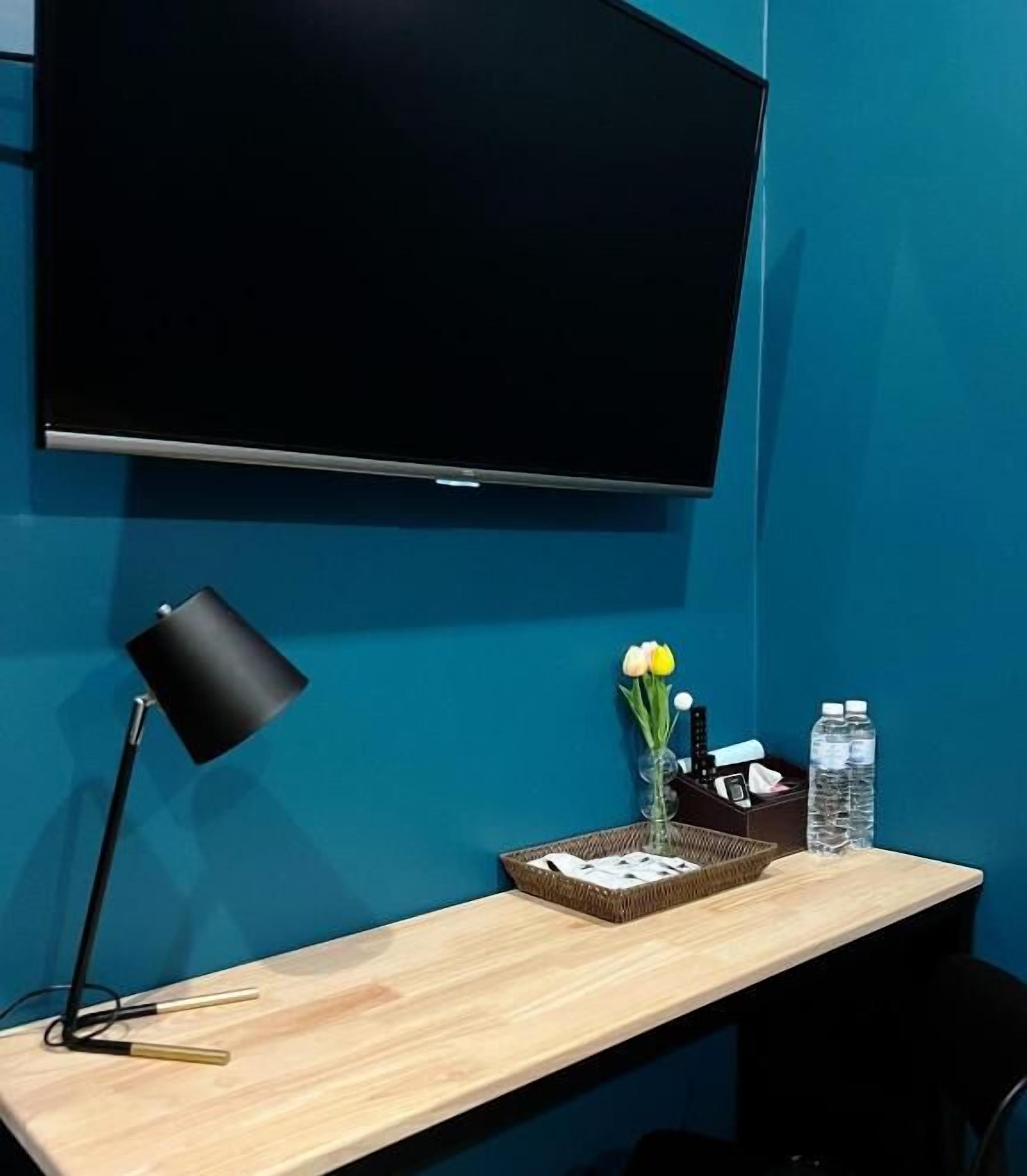 Ein Zimmer mit einem großen Flachbildfernseher an der Wand, einem Schreibtisch mit Lampe, einer Vase mit Blumen, einem Korb und zwei Wasserflaschen.