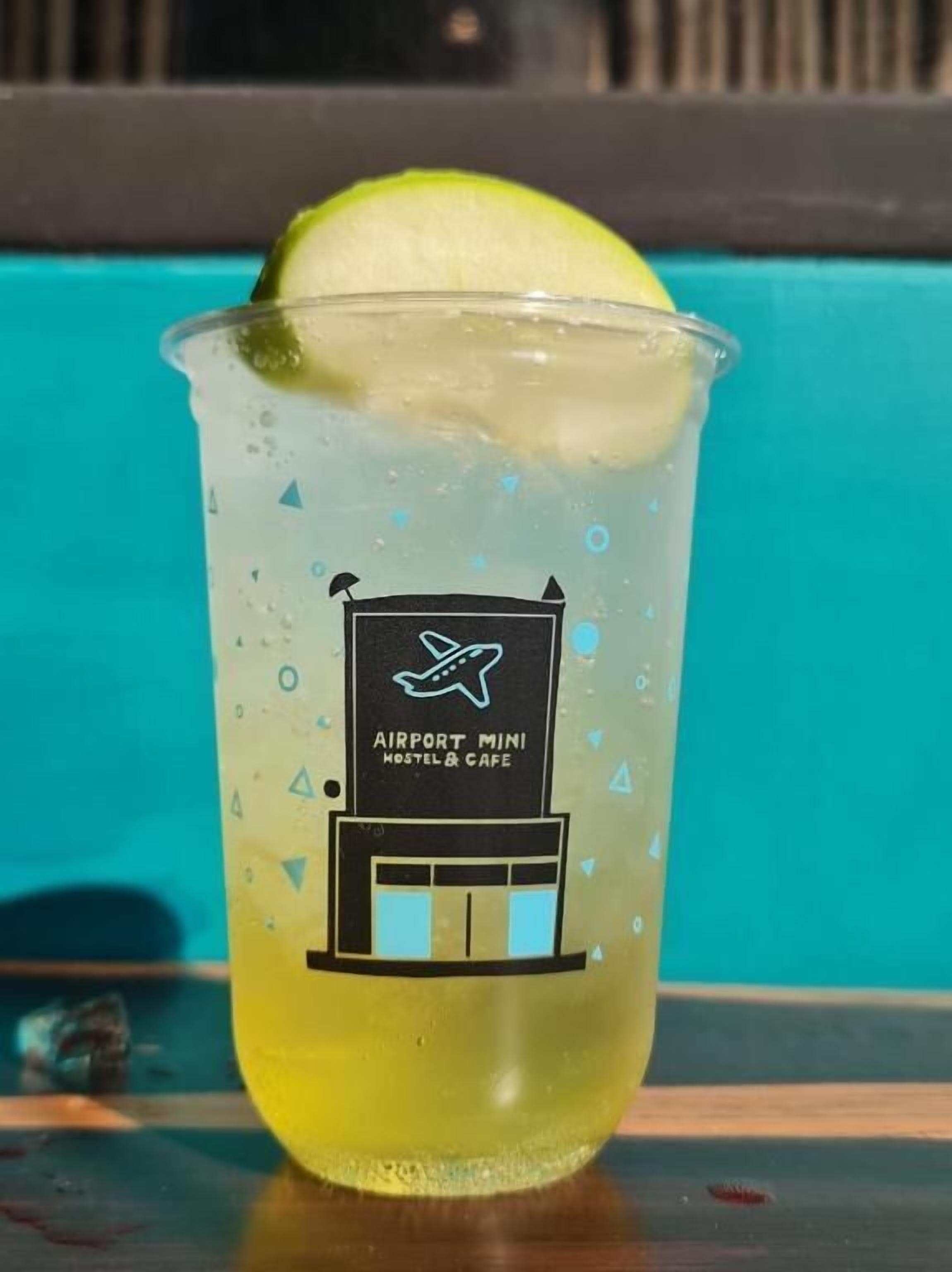 A clear plastic cup with a lime wedge, labeled "AIRPORT MINI HOSTEL & CAFE" with a building and airplane icon.