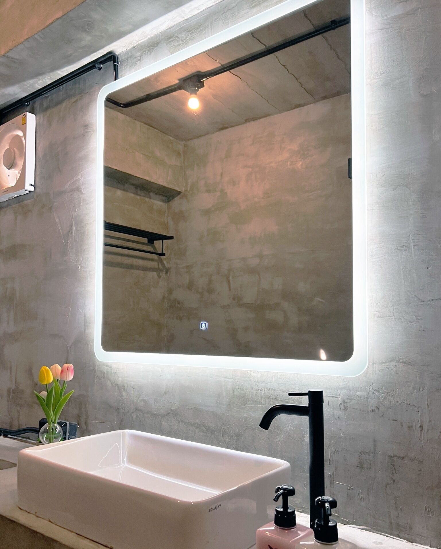 Un baño con lavamanos blanco, grifo negro y un espejo grande con luz. Hay flores en un jarrón sobre la encimera.