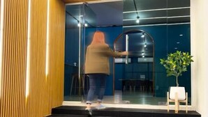 Une personne ouvre une porte coulissante en verre à l’entrée d’un bâtiment au design moderne, caractérisé par des lambris en bois et un intérieur sombre.