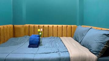 Una cama con una colcha azul, una manta color beige y un pequeño jarrón con flores.