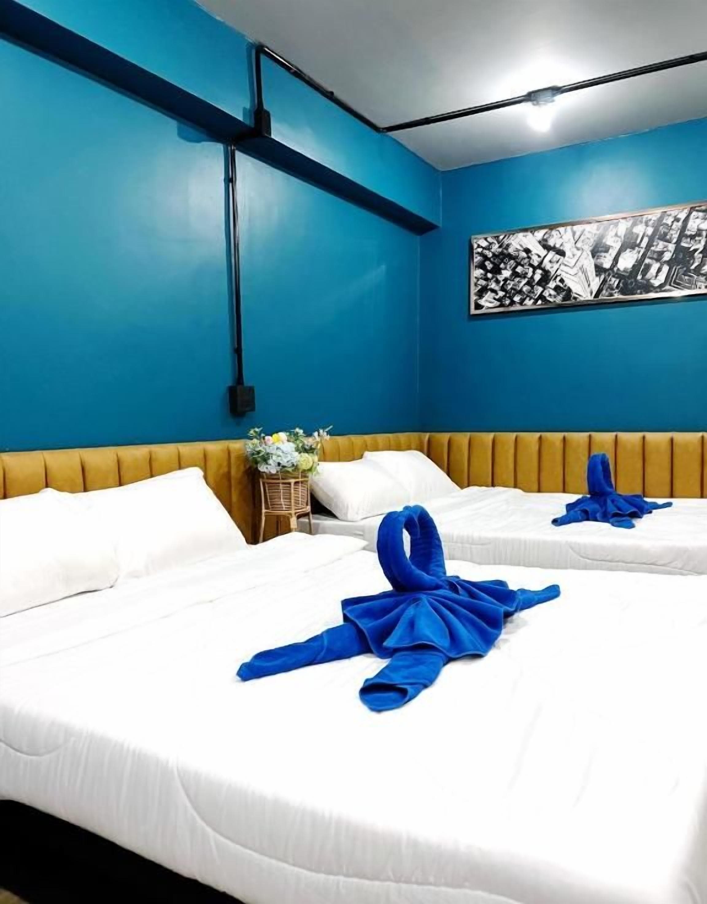 Une chambre d’hôtel avec deux lits, des murs bleus, une tête de lit en bois et une œuvre d’art représentant un paysage urbain en noir et blanc.