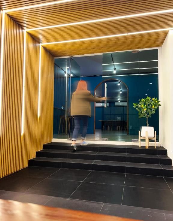 Une personne ouvre une porte coulissante en verre à l’entrée d’un bâtiment au design moderne, caractérisé par des lambris en bois et un intérieur sombre.