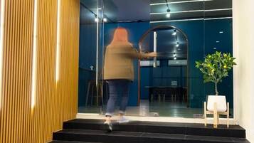 Une personne ouvre une porte coulissante en verre à l’entrée d’un bâtiment au design moderne, caractérisé par des lambris en bois et un intérieur sombre.