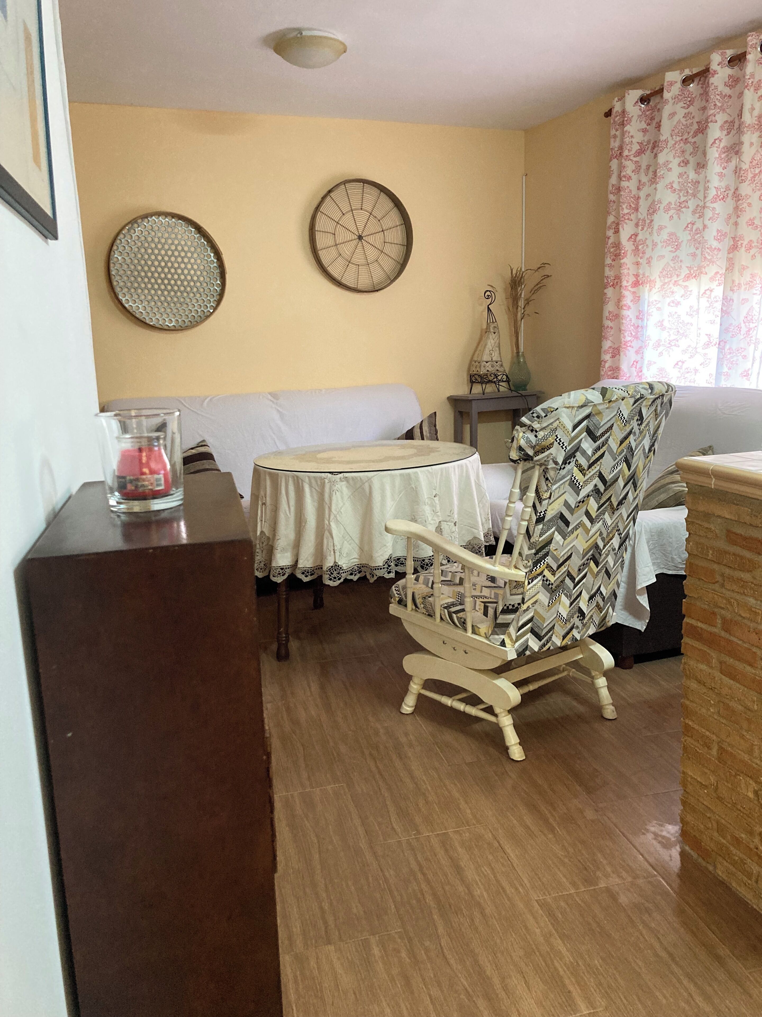 Una habitación con piso de madera, un sillón blanco con funda estampada, una mesita con mantel de encaje y un ventilador de pared.