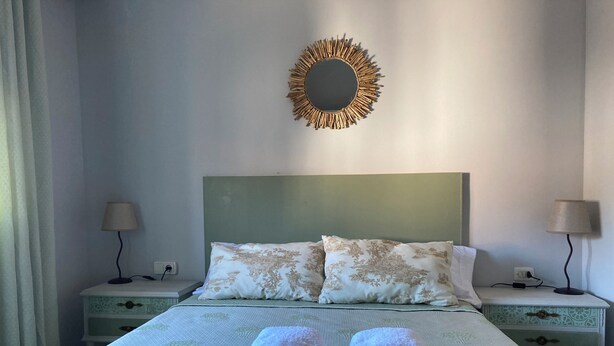 Una cama bien tendida con dos almohadas, una mesita de noche con lámpara y un espejo montado en la pared.