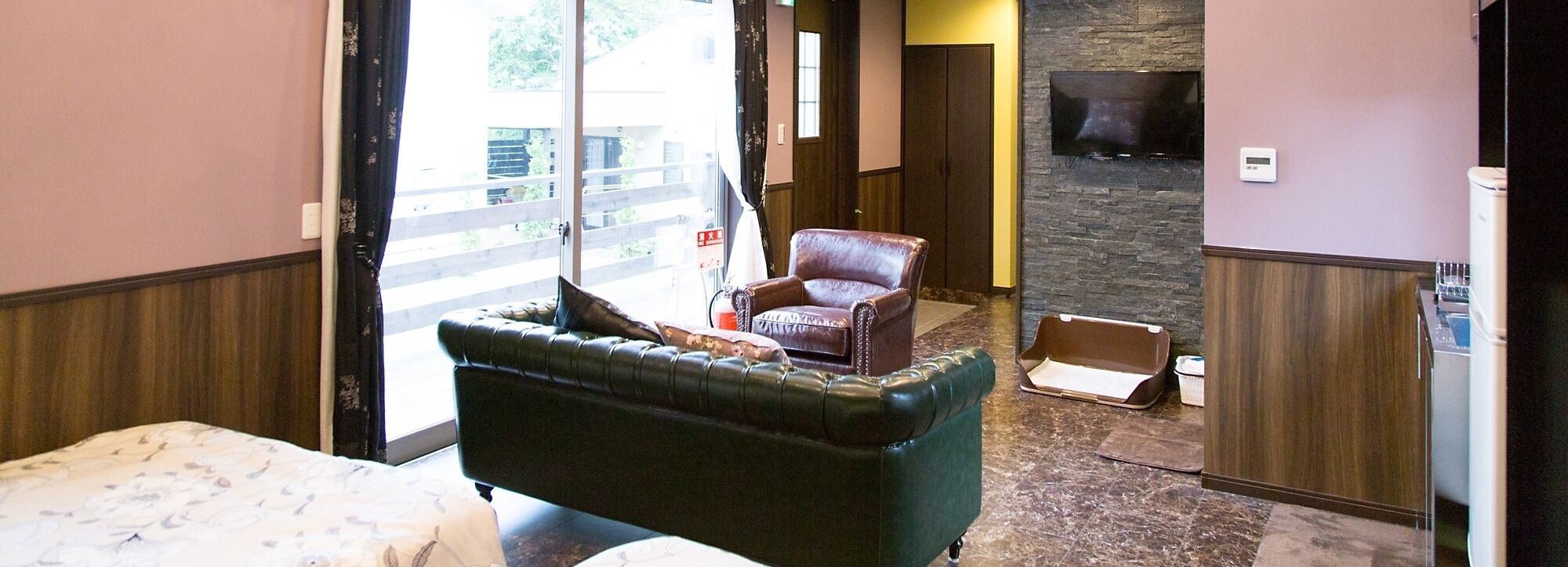 A hotel room with a green upholstered sofa, a leather armchair, a flat-screen TV mounted on a stone wall, and a wooden floor.