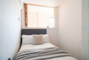 A small, neatly made bed with a grey headboard and a window above it. - Stunning 2 Bedroom Manchester Apartment w Balcony (Manchester)