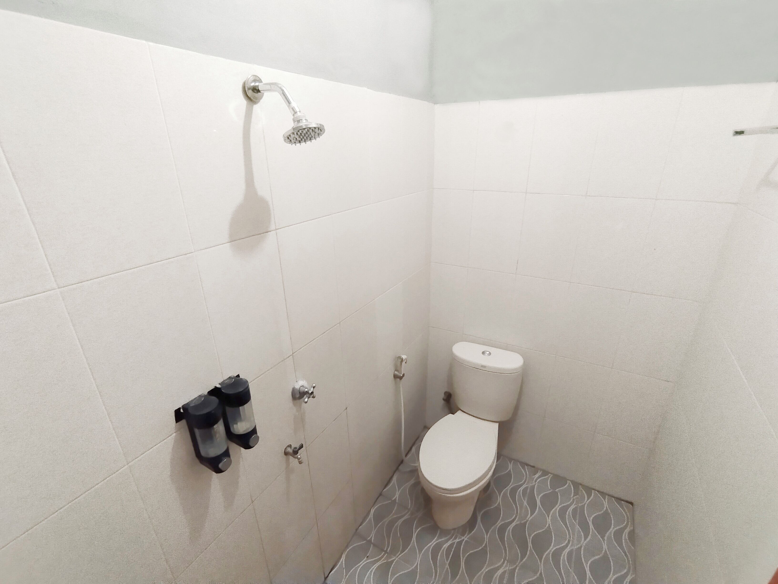 Um banheiro pequeno com azulejos, chuveiro, vaso sanitário e dois dispensadores pretos na parede.