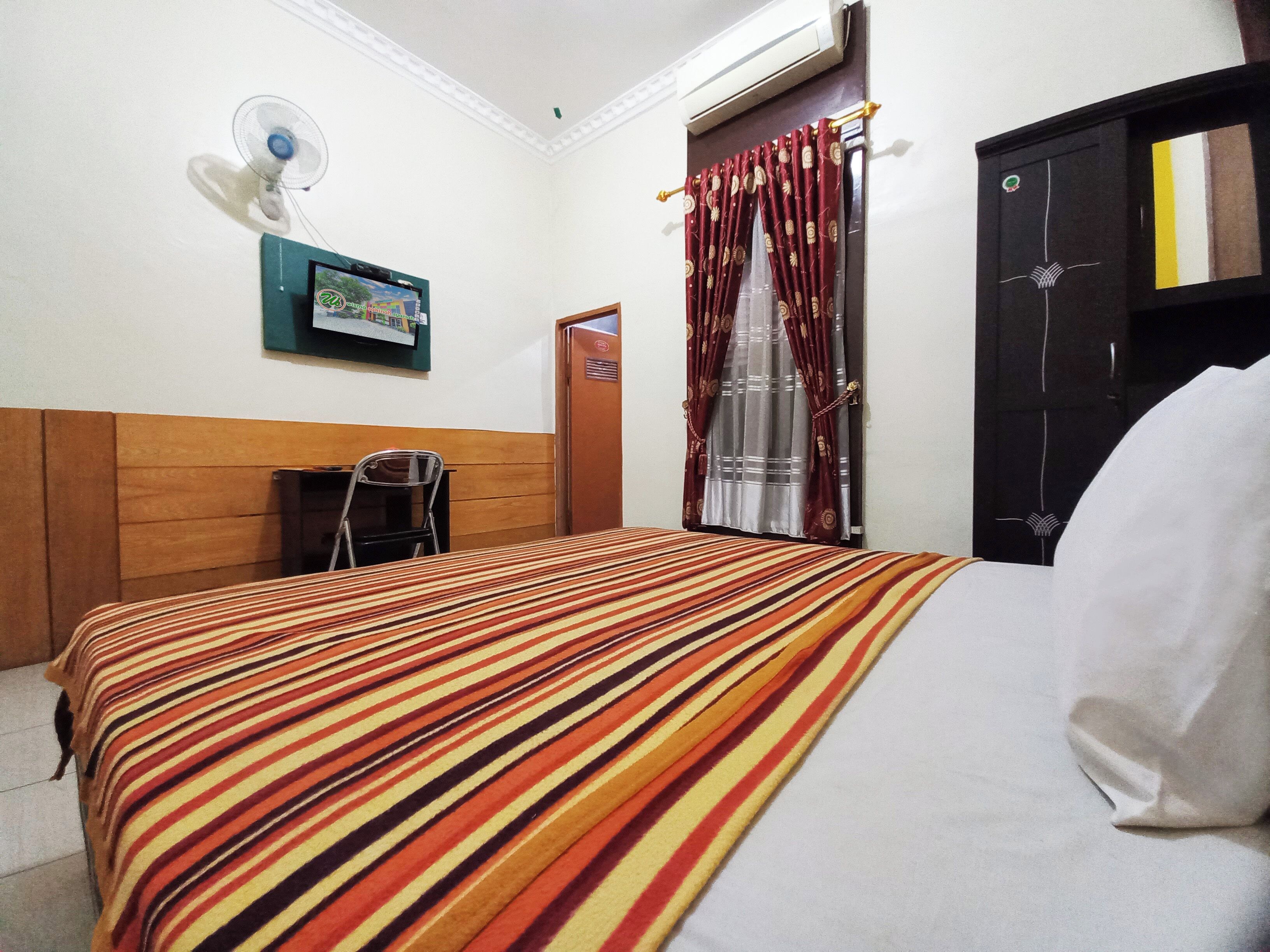 A hotel room with a striped bedspread, a wall-mounted fan, a door with a curtain, and a small table with a chair.