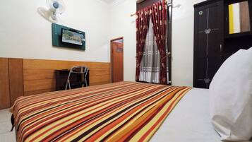 A hotel room with a striped bedspread, a wall-mounted fan, a door with a curtain, and a small table with a chair.