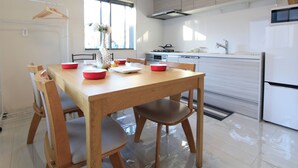 A modern kitchen with white cabinets, a stainless steel range hood, and a built-in oven. There is a dining area with a wooden table and chairs, and a window providing natural light.