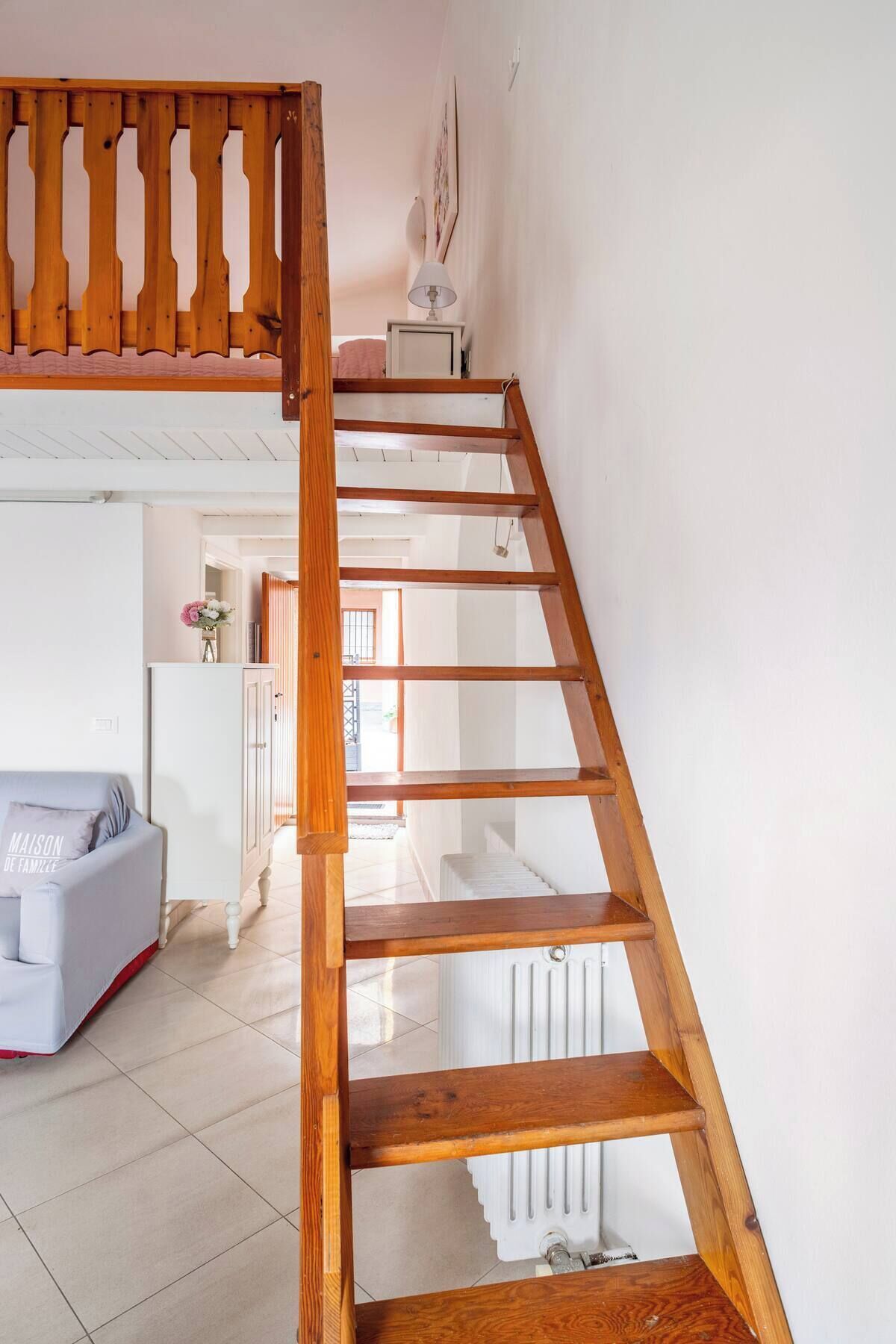 Una escalera de madera lleva a un dormitorio en entrepiso con techo blanco y vista a una sala de estar con sofá y radiador.