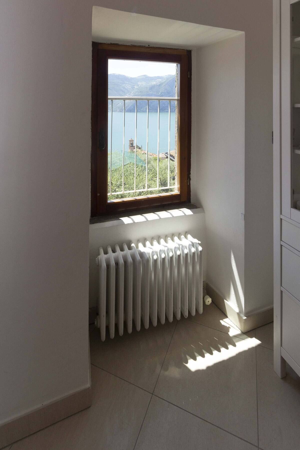 Een kamer met een raam dat uitkijkt over een meer en bergen, een radiator en een hoekkast.